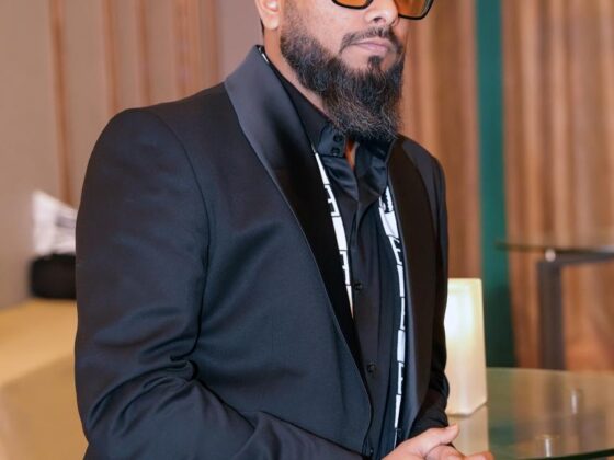 A man with a neatly trimmed beard and short styled hair stands indoors wearing a black suit, black shirt, and amber-tinted rectangular glasses. He has a black-and-white patterned scarf draped around his neck and clasps his hands in front of him. The background features warm wooden wall panels, soft lighting, and a modern high table, creating a professional event or business conference setting.